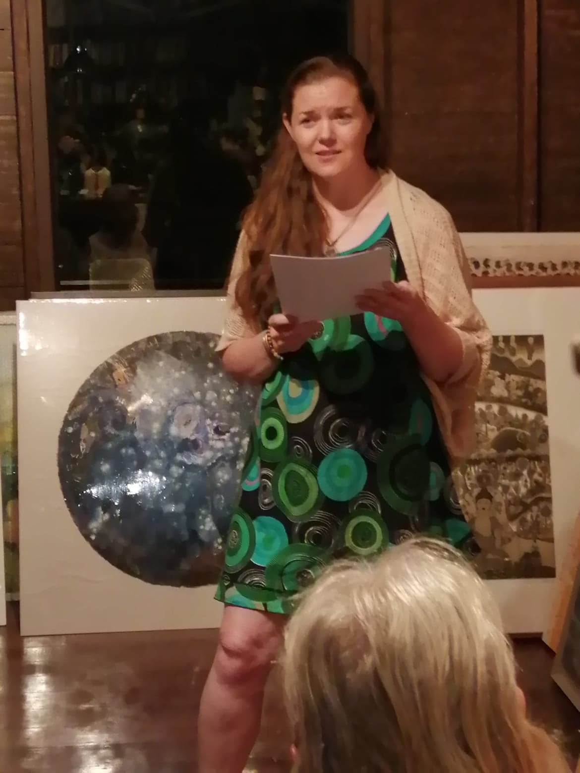 A white woman stands at the front of a cafe holding a piece of paper and speaking ardently as she looks off to the side.