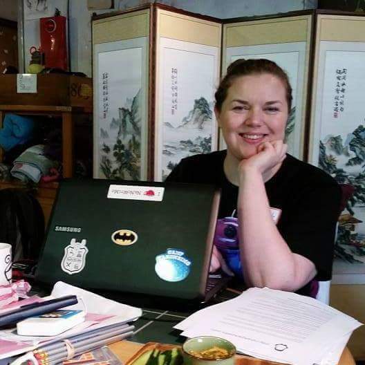 A white woman sits at a table with a laptop in front of her, smiling with her elbow on the table and chin in her hand.